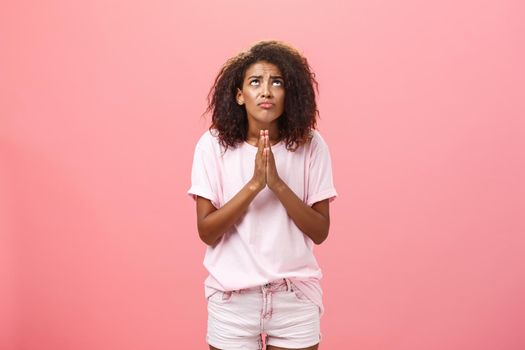 Studio shot of unhappy miserable and hopeless cute african american female holding hands in pray near chest looking up with serious-looking concerned expression making wish to god over pink wall. Lifestyle.