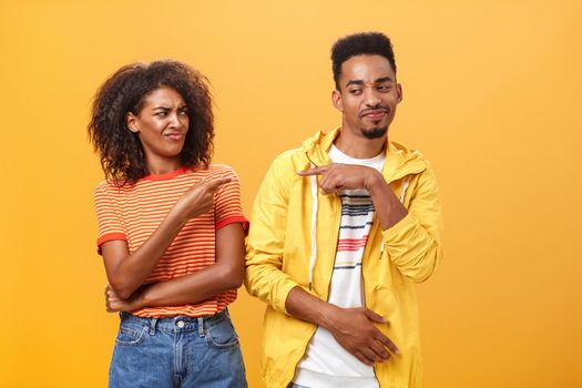 Two best friends fooling around having fun pointing at each other with cute joyful smile as if blaming each other playfully. Cute female looking at boyfriend standing together over orange wall.