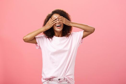 Portrait of charming joyful dark-skinned playful woman with curly hair in t-shirt closing eyes and counting ten with broad happy smile playing hide n seek or waiting for surprise over pink background. Lifestyle.