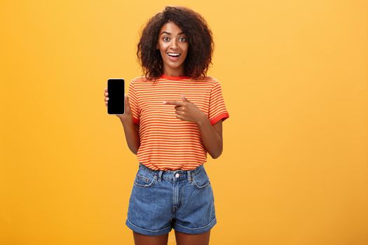 Cute african american stylish woman showing friend new app in smartphone pointing at cellphone screen with index finger smiling thrilled and happy delighted with cool device purchase over orange wall. Lifestyle.