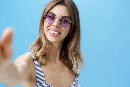 Charming woman with cute gapped teeth in sunglasses relaxing on vacation taking beautiful shots pulling hand towards camera holding it to take selfie smiling broadly over blue background. Lifestyle.