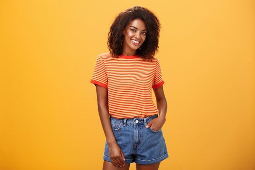 Ready to travel world. Energetic confident and attractive dark-skinned woman with curly hair holding hand in pocket of denim shorts smiling joyfully posing over orange background carefree and friendly.