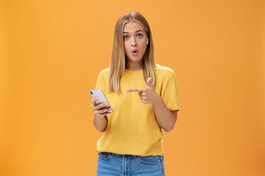 Wman impressed by wireless earphones connecting to smartphone themselves pointing at device screen questioned and surprised folding lips looking intrigued at camera wearing earbuds over orange wall. Technology and advertisement concept