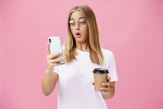 Woman drinking coffee being shocked by received message reacting on stunning news folding lips gasping looking astonished and impressed at smartphone screen holding paper cup, posing over pink wall. Technology concept