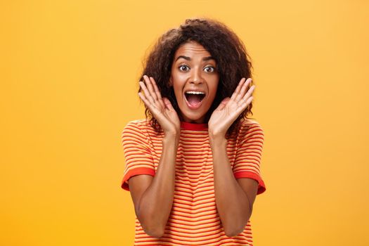 Waist-up shot of impressed excited talkative dark-skinned woman with curly hair and perfect skin yelling from happiness and amazement shaking palms near face cheering for awesome sale over orange wall. Lifestyle.