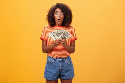 Woman shocked finding lots of cash in safe. Portrait of surprised speechless good-looking dark-skinned female with curly haircut folding lips gasping holding money posing over orange background.