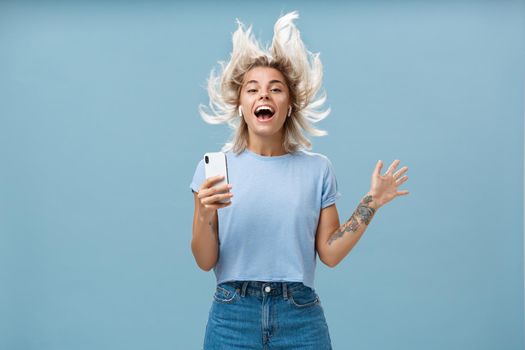 Expressing happiness with help of great tunes. Joyful amused and happy good-looking young female student jumping having fun listenign music in wireless earbuds, holding smartphone over blue wall.