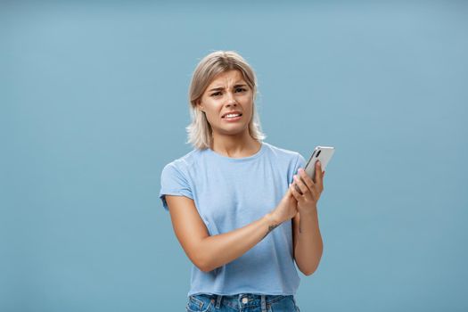 Woman cannot stand when mom shouts during phone call. Intense displeased young european blonde with tanned skin clenching teeth and frowning covering microphone of smartphone over blue wall.