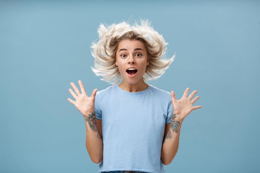 Waist-up shot of impressed surprised attractive and cute girl with blonde hair floating in air and tattooed arm jumping with spread raised palms and opened mouth over blue background. Emotions concept