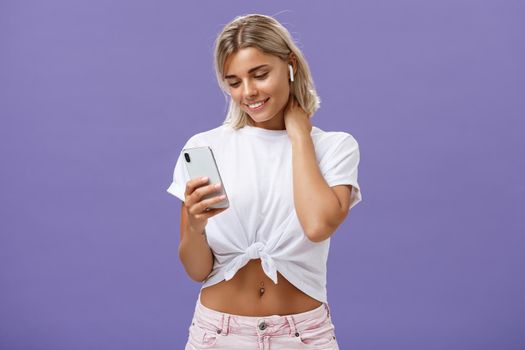 Woman receiving warm romantic message being delighted and touched feeling love gazing at smartphone screen wearing wireless earphones touching neck gently being sensitive and feminine.