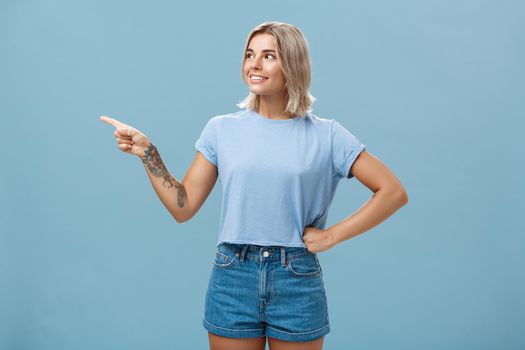 Studio shot of intrigued charming young european female with tanned skin and blond hair holding hand on waist in confident pose looking and pointing left with interest and curiosity. Copy space