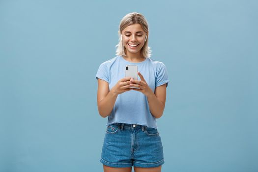 Girl having funny convesation, speaking with friend via video messages in wireless earphones holding smartphone in both hands smiling happily at device screen being entertained and amused. Emotions and technology concept