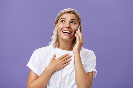 Lifestyle. Waist-up shot of thankful charming blonde female student with tanned skin and blond hair holding hand on breast receiving delightful warm call from loving friend holding smartphone near ear gazing up.