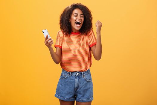 Portrait of ambitious happy young african american girl yelling from happiness and triumph clenching fist in joy and celebration feeling excited and relieved holding smartphone over orange wall. Lifestyle.