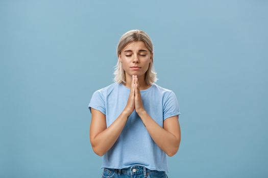 Lifestyle. Good-looking kind and faithful european girl with blond hair smiling cute and tender holding hands in pray while making wish hopefully believing god hearing her prayers with closed eyes over blue wall.