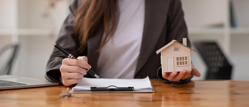 Close up businesswoman realtor taking notes and holding house model, sitting at desk with paper house model and keys, female real estate agent manager, preparing documents, mortgage and property