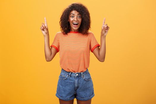 Charmed and impressed african american stylish woman seeing incredible and awesome item on sale raising hands and pointing up with broad grin being excited of new purchase over orange wall. Emotions and advertisement concept