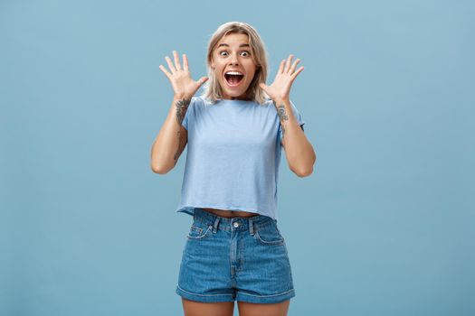 You gotta listen this. Thrilled amused and amazed impressed good-looking young european female with blond hair gasping holding breath and smiling broadly raising palms from surprise over blue wall. Emotions concept