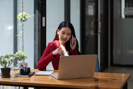 Smiling business woman at work talking on phone, asian woman sitting and working financial with laptop computer.