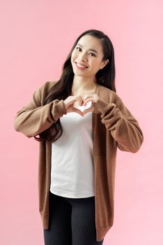 Young woman standing isolated on pink background showing heart shape hand gesture looking camera smiling cheerful