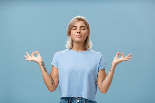 Listening to inner self during meditation. Calm happy and relaxed attractive tanned blonde female with tattoos on arms standing in lotus pose over blue wall with zen gesture closed eyes doing yoga.