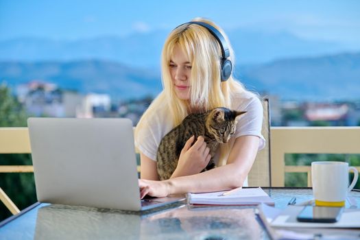 Female student studies at home online using laptop. Teenager sitting on outdoor balcony with pet cat in headphones, school notebooks, looking at screen. E-learning, modern technologies in education