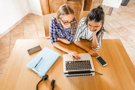 Two diverse girls view from top working or studying at laptop computer sitting at home table with note book, pen, headphone and smartphone. Gen z students using wi-fi to Attend online classes together