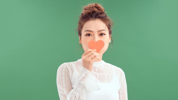 Waist-up portrait of pretty Asian woman looking away with toothy smile while holding Valentines Day card in hands, white background