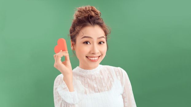 Waist-up portrait of pretty Asian woman looking away with toothy smile while holding Valentines Day card in hands, white background