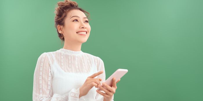 Photo of cheerful cute beautiful young woman chatting by mobile phone isolated over green wall background. Looking aside.