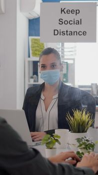 Freelancer with face mask working at financial graphics on pc sitting in business office. Coworkers talking in background about marketing mantain social distancing to prevent infection with covid19