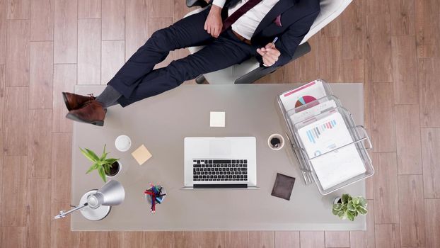 Top view of businessman in suit staying relaxed with feet on office desk analyzing financial graphs on laptop. Executive manager working in startup company office at management investments