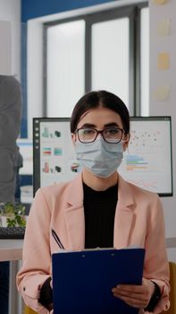 Entrepreneur sitting in new normal office looking into camera while writing lifestyle information during online videocall meeting. Businesswoman wearing face mask to avoid infection with covid19