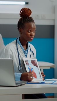 Young medic holding tablet with medical cardiogram sitting at desk in office. Elder patient looking at screen gadget with heart figure while doctor explaining cardiac disease