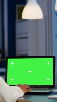 Back view of black business woman looking at laptop with greenscreen, green mockup, chroma key desktop sitting at desk in start up business office overworking. Freelancer watching at isolated display