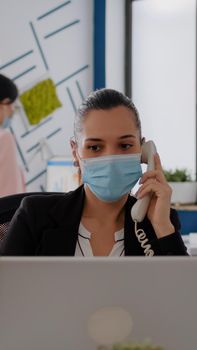 Entrepreneur with protective face mask discussing on landline while sitting at startup office desk in front of computer. Caucasian female working at business meeting during coronavirus global pandemic
