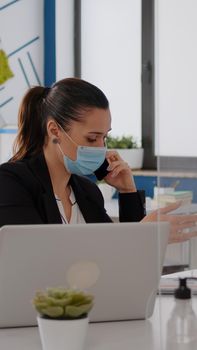 Woman with protective face mask working on laptop computer in company office checking statistics while talking on phone.Team workers respecting social distancing to avoid infection with covid19 virus
