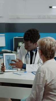 Doctor holding tablet explaining bones fracture recovery to old patient in healthcare office. Elderly woman looking at digital device screen for chiropractic annual medical checkup