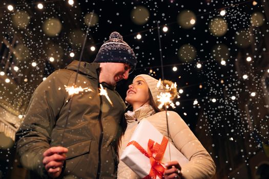 Close up portrait of young happy couple standing on the street hugging holding sparklers. Young couple in love holds sparklers in their hands. Two people celebrate New Year on the street.