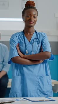 Portrait of medical assistant wearing uniform standing in cabinet at healthcare facility. Woman working as nurse smiling looking at camera in doctors office with medic and patient