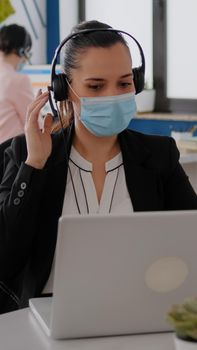 Close up of businesswoman with face mask wearing headset talking into microphone while typing marketing statistics on laptop computer. Freelancer sitting at desk in office during covid19 pandemic