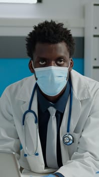 Close up of young medic with face mask in medical cabinet using modern laptop on desk. Doctor sitting with digital device typing on keyboard while wearing stethoscope for healthcare