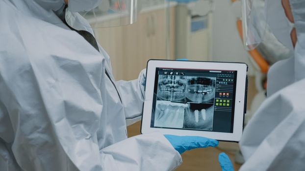 Close up of teeth radiography scan on modern tablet while dental specialist holding device and assistant looking at screen. Stomatologists wearing ppe suits examining oral x ray