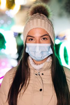 Close up portrait of Caucasian female in medical face mask standing in decorated xmas city. Crowd people sick covid-19. Epidemic coronavirus. Pandemic flu corona virus