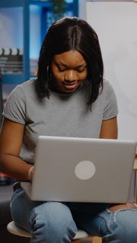 Black young artist typing on laptop for art project while sitting in workshop space at home. African american creative woman using device and technology for masterpiece and craft concept