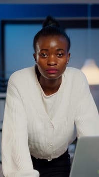 Focused tired business woman looking at camera after reading tasks on laptop standing near desk in start-up company late at night. Employee doing overtime for job respecting deadline