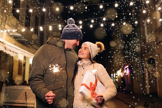 Lovers travelling to Europe at the height of Christmas holidays. Sweethearts in the centre of city. Romantic couple in love with gift box hugging and burning the sparklers