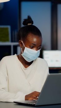 African woman with face mask reading emails late at night to respect deadline of project working in new normal business office, analysing documents, making strategy overtime during global pandemic