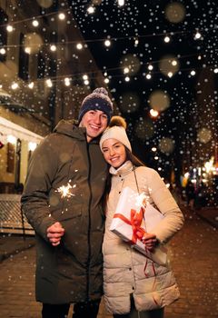 Romantic couple in love with gift box hugging and burning the sparklers. Lovers travelling to Europe at the height of Christmas holidays. Sweethearts in the centre of city.