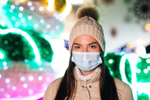 Epidemic coronavirus. Pandemic flu corona virus. Close up portrait of Caucasian young beautiful female in medical mask standing in decorated xmas city. Crowd people sick covid-19.